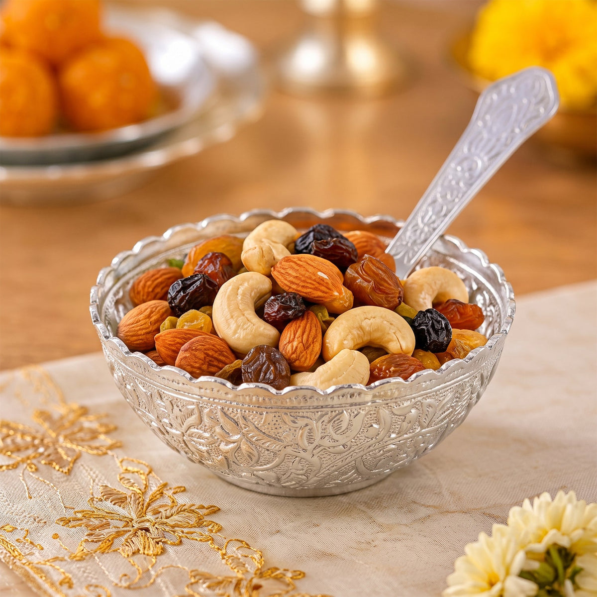Silver finish floral serving bowl filled with mixed nuts and dried fruit, with a spoon on a patterned cloth.