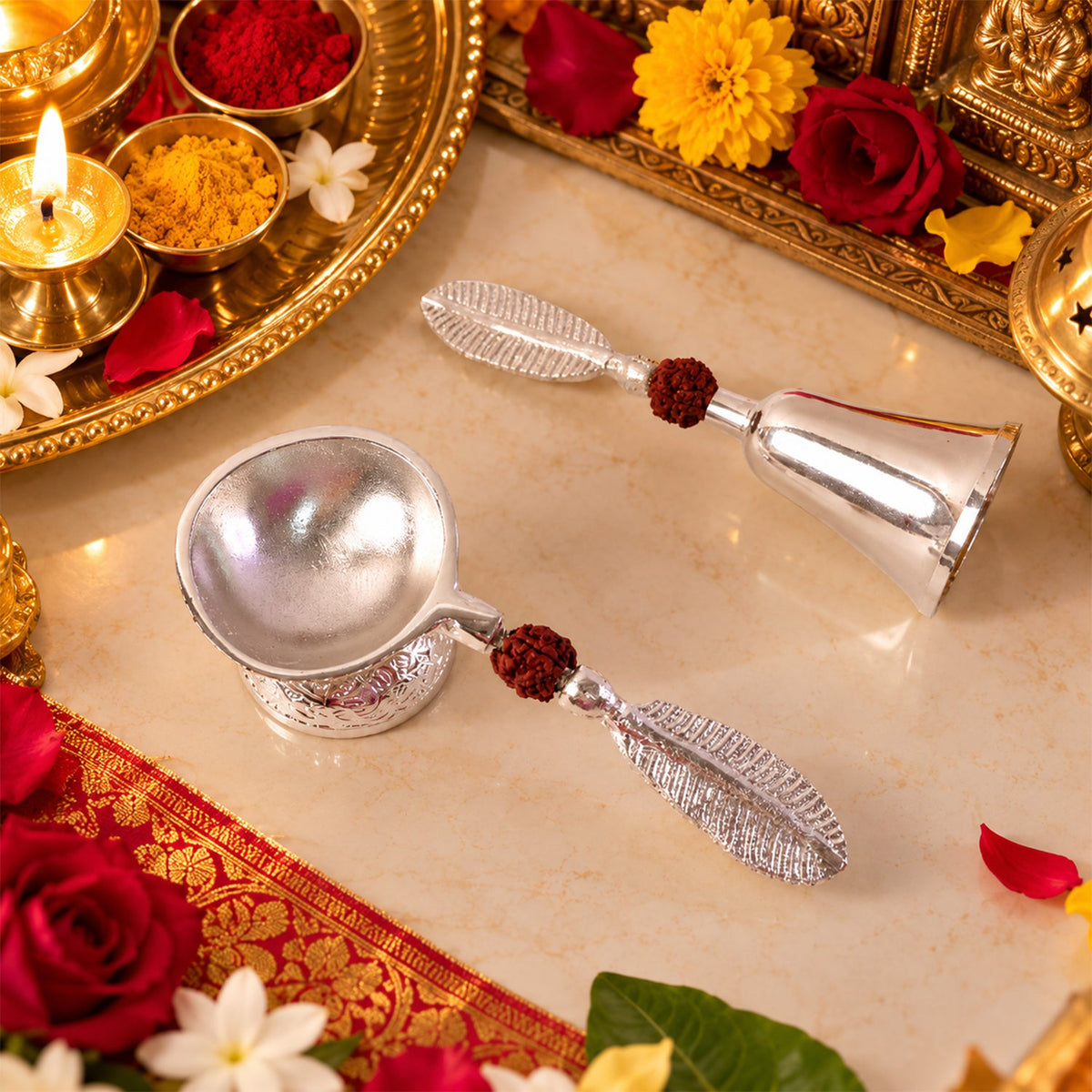 A silver finish pooja bell and diya set with leaf handles and rudraksha beads, surrounded by colorful flowers and religious