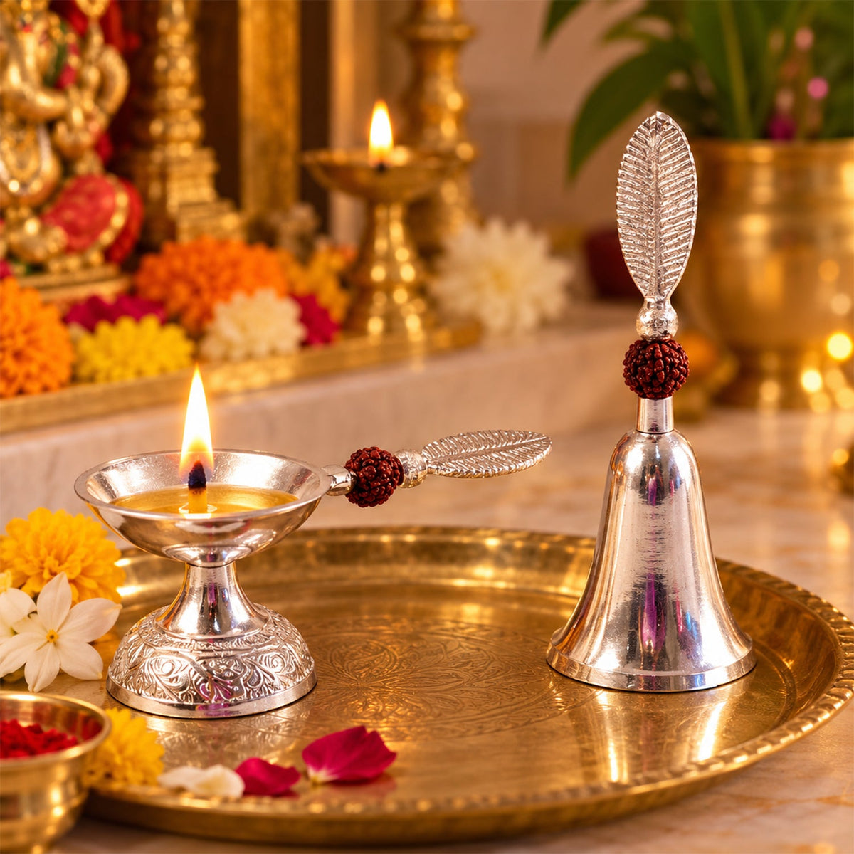 A silver finish pooja bell and lit diya set with leaf handles and rudraksha beads, on a golden brass tray, surrounded by