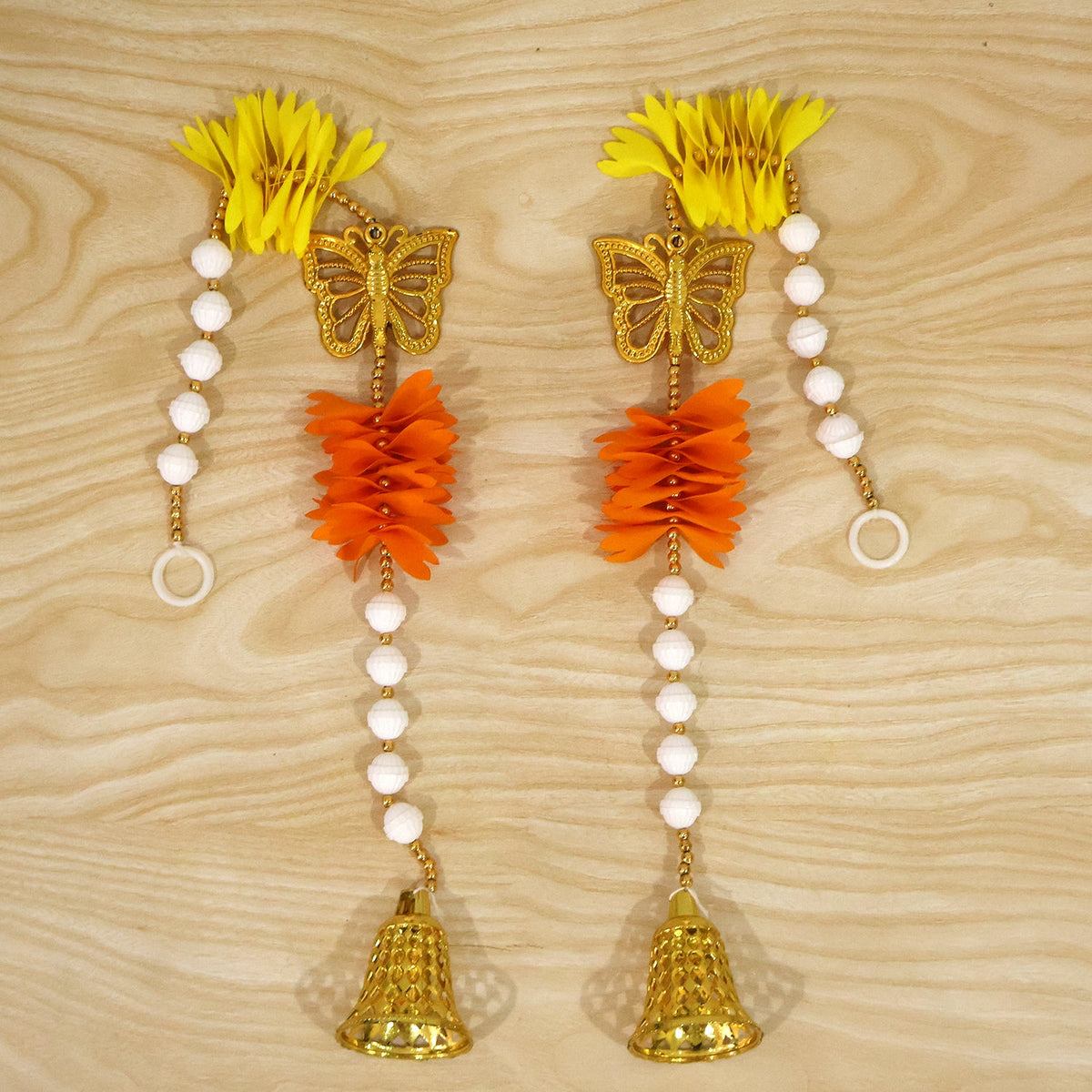 A pair of decorative door hangings, each with yellow and orange paper flowers, a golden butterfly, and a bell, displayed on