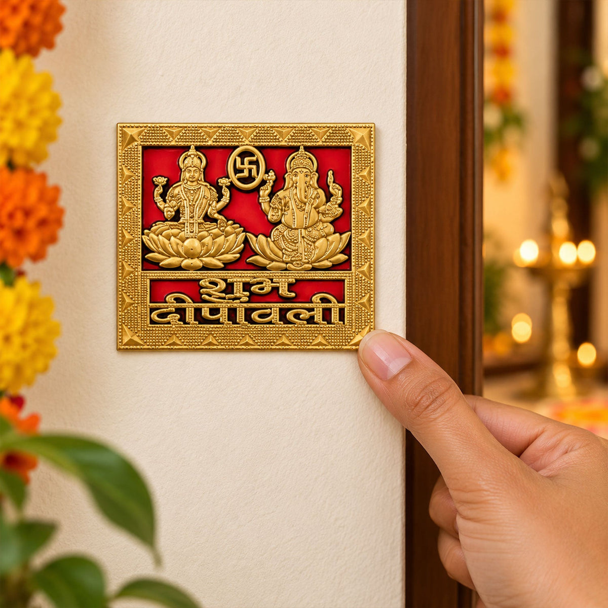 A hand applies a square golden sticker with embossed Lakshmi and Ganesha figures on a red background to a wall.