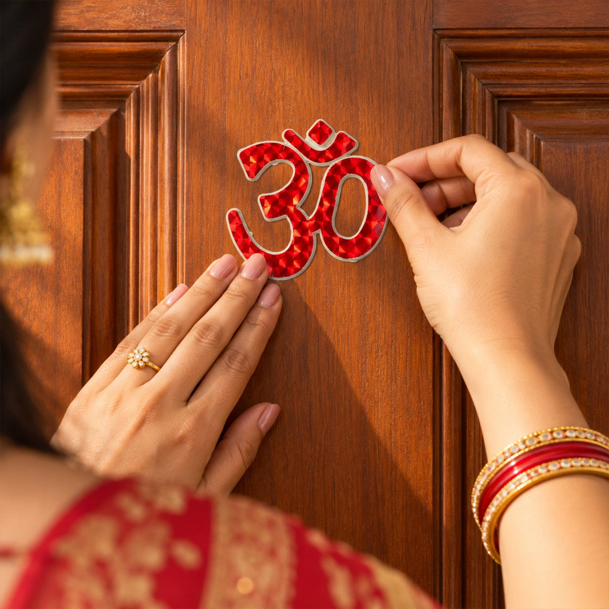 Hands carefully place a red Om symbol sticker onto a rich brown wooden door, adorned with bangles and a ring.