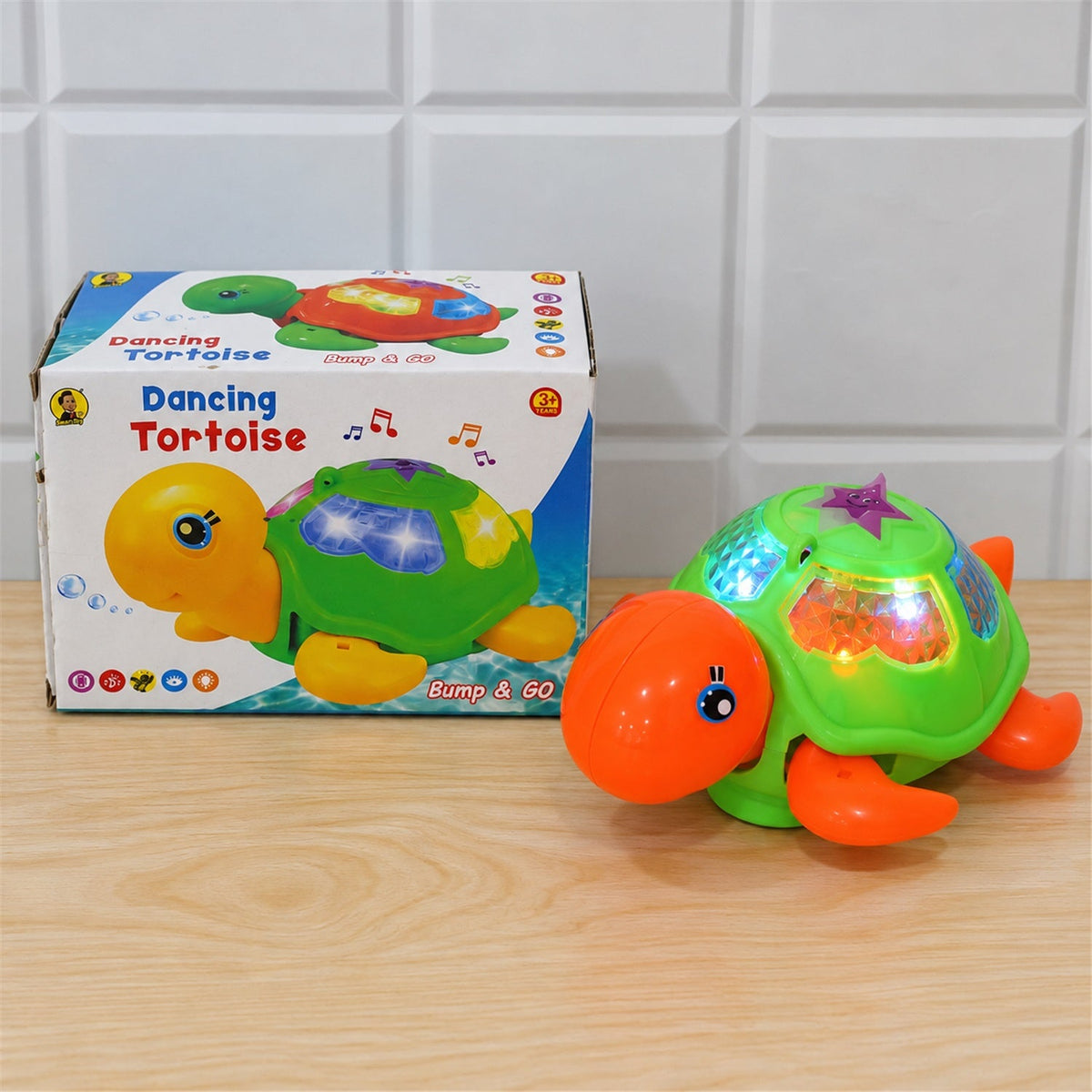The green and orange tortoise toy with flashing lights sits on a wooden surface beside its