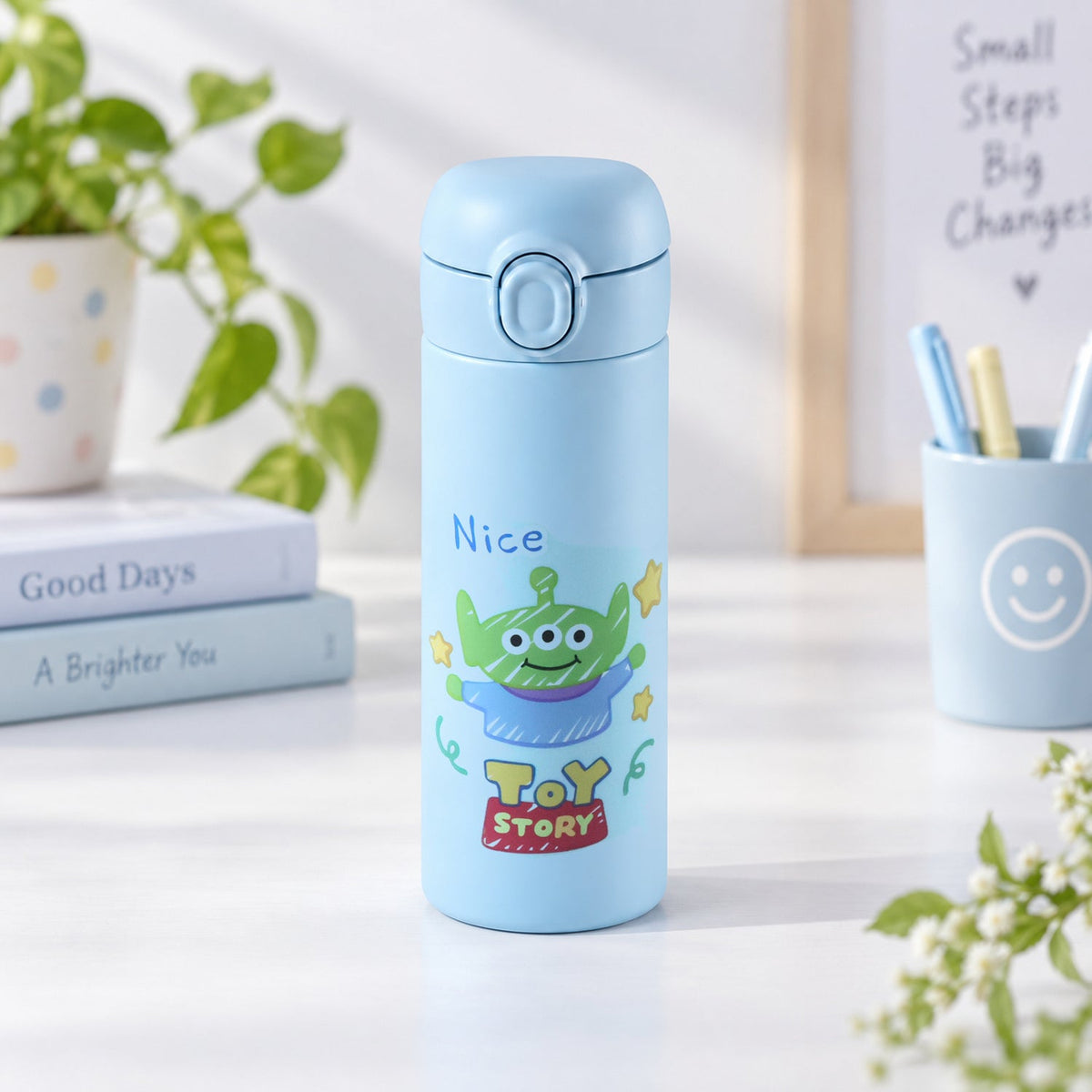 A light blue water bottle with a cartoon alien design stands on a desk beside books and a pen holder.