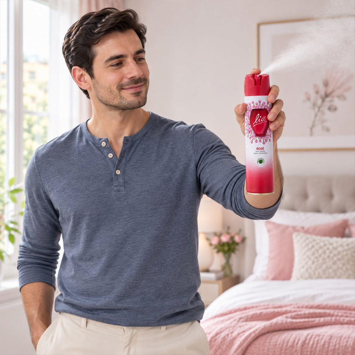 Man in a blue long-sleeve shirt spraying the red and white room freshener in a bedroom with pink bedding.