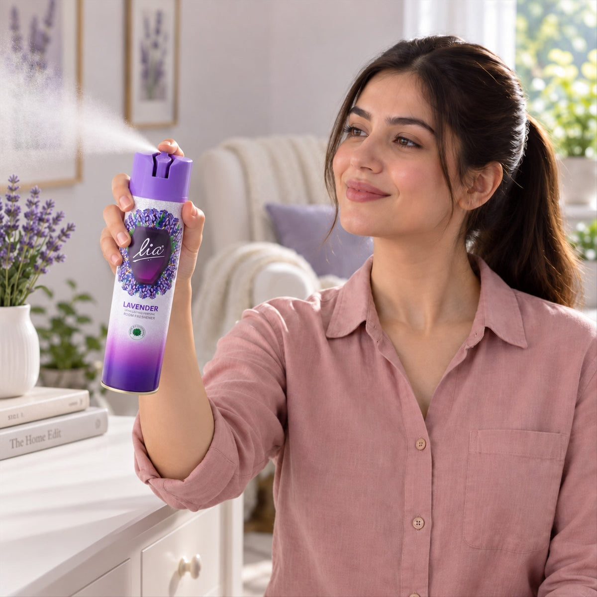A woman in a pink shirt sprays the room freshener, looking right, in a bright room.