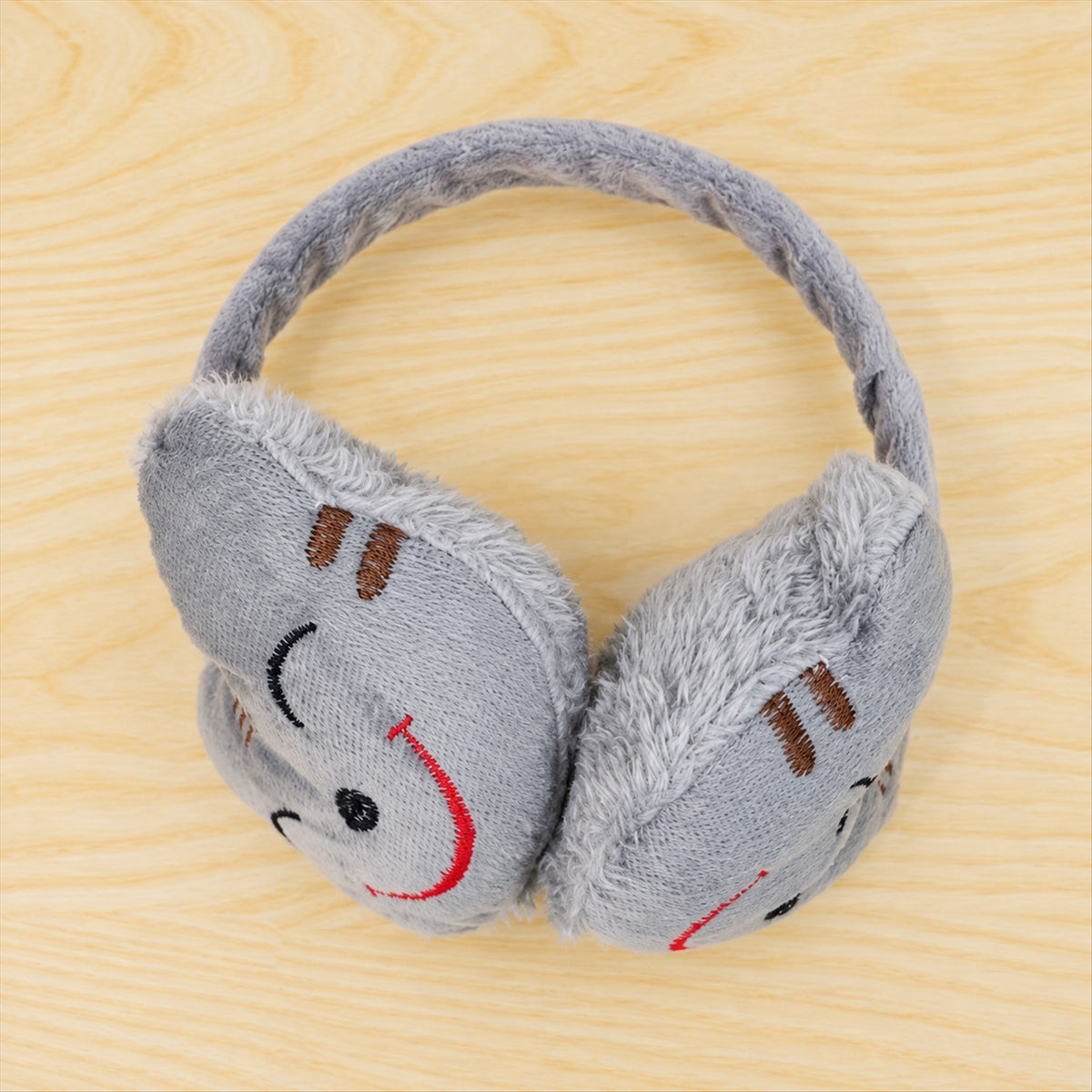 Grey plush earmuffs with a red embroidered smiley face on a light wooden surface.