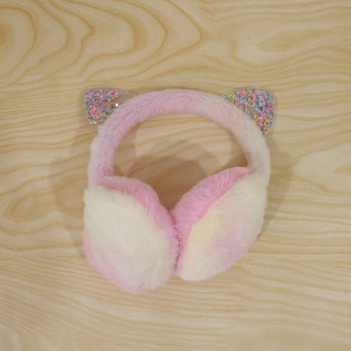 The pink and cream fluffy earmuffs with clear, star-filled cat ears are placed on a light wooden surface.