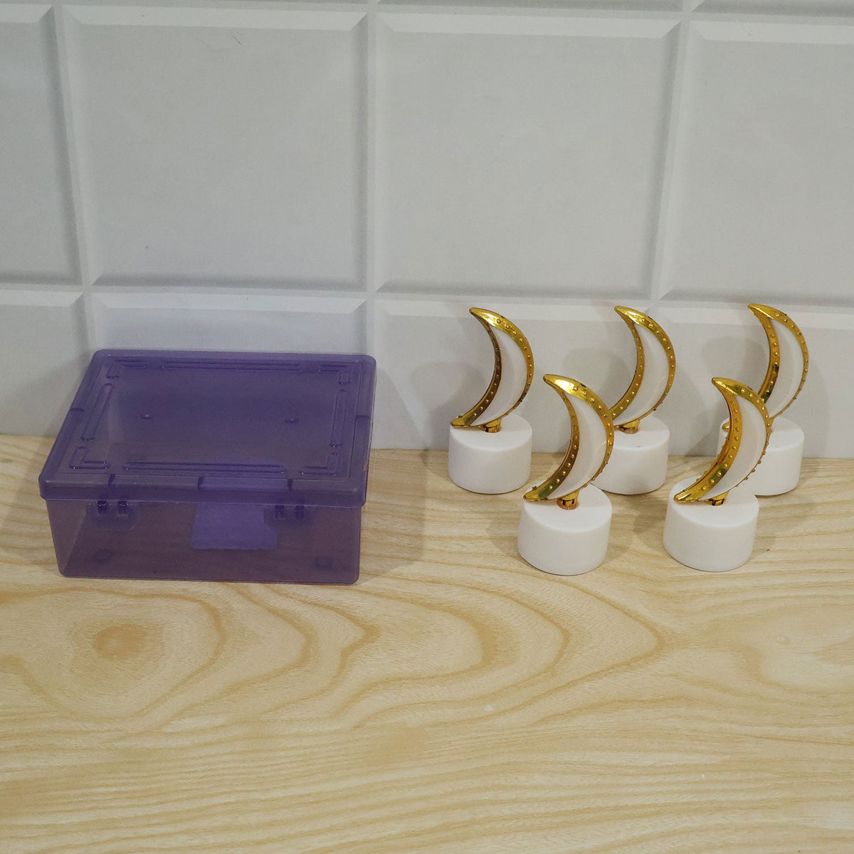 Five unlit moon-shaped LED tealights with white bases and golden tops, placed next to a translucent purple storage box on a