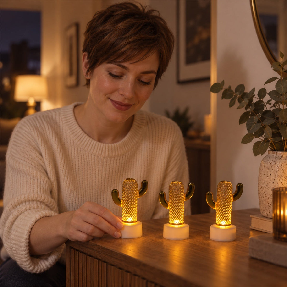 A woman with short brown hair smiles while touching one of three glowing gold cactus lights on a wooden dresser.