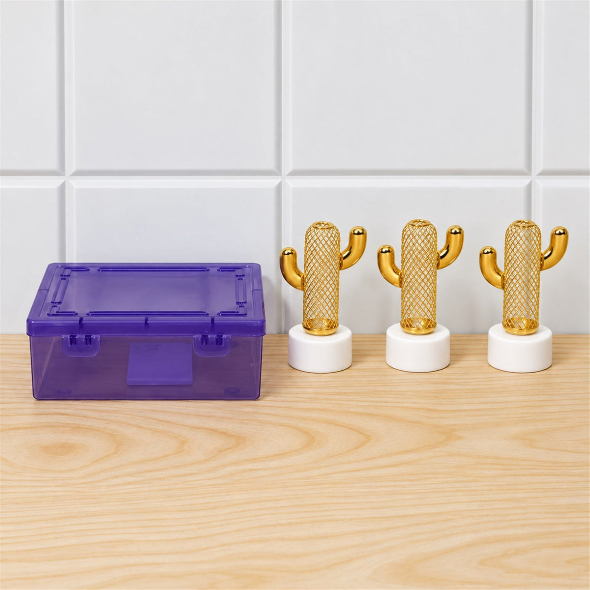 Three gold cactus lights with white bases are next to a translucent purple storage box on a light wooden surface.