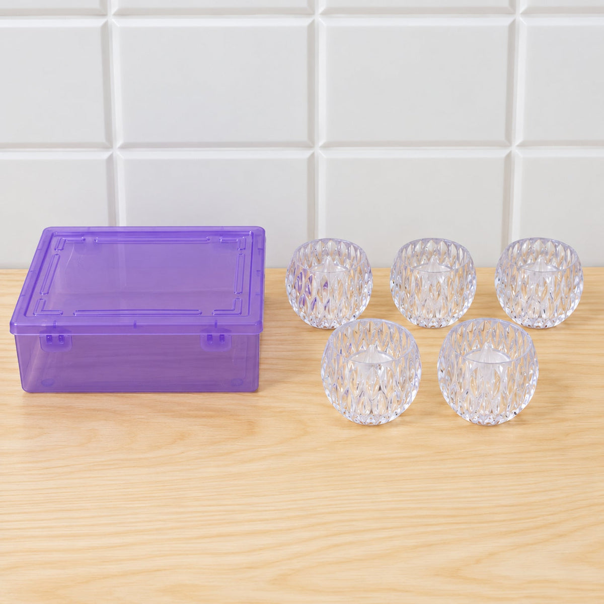 Four empty crystal tealight holders and one with a light sit beside a purple plastic storage box on wood.