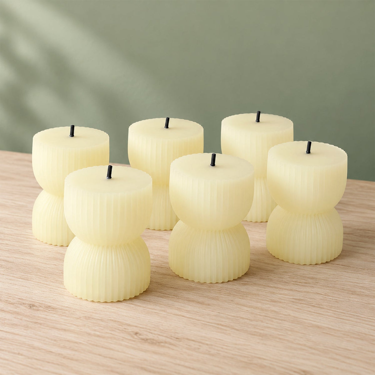 Six unlit cream-colored ribbed LED candles with black wicks arranged on a light wooden surface against a green wall.