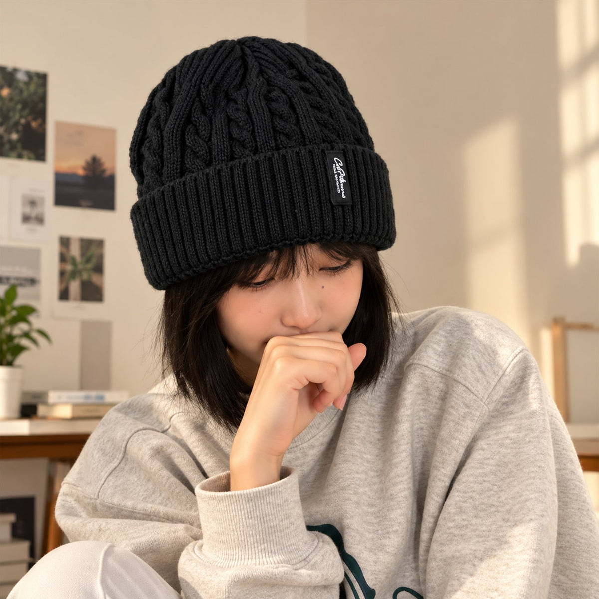 A young woman wearing a black cable knit beanie, looking down thoughtfully in a cozy indoor setting.