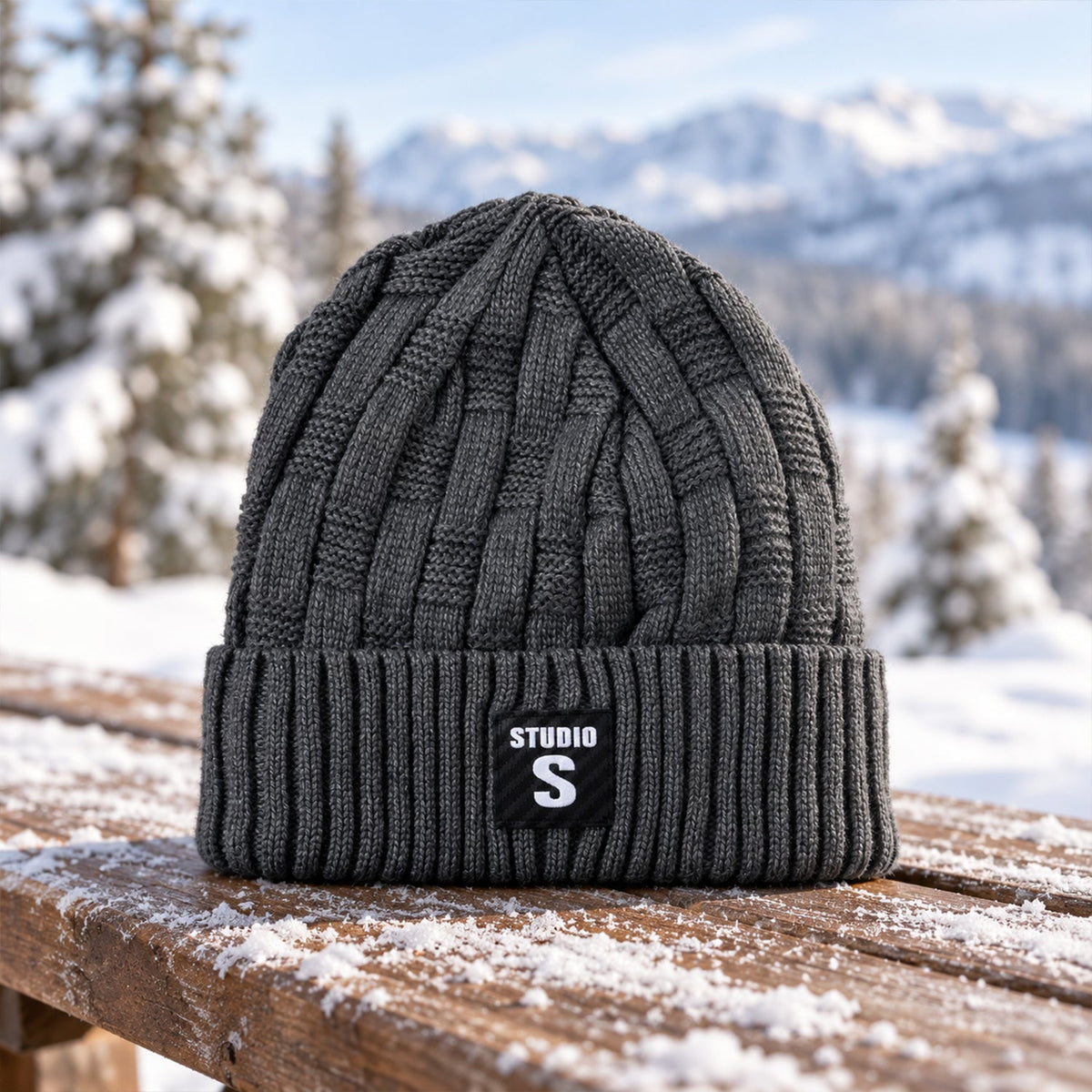 A dark grey cable knit beanie with a ribbed cuff and a black label with "STUDIO S" in white, resting on a snow-dusted wooden