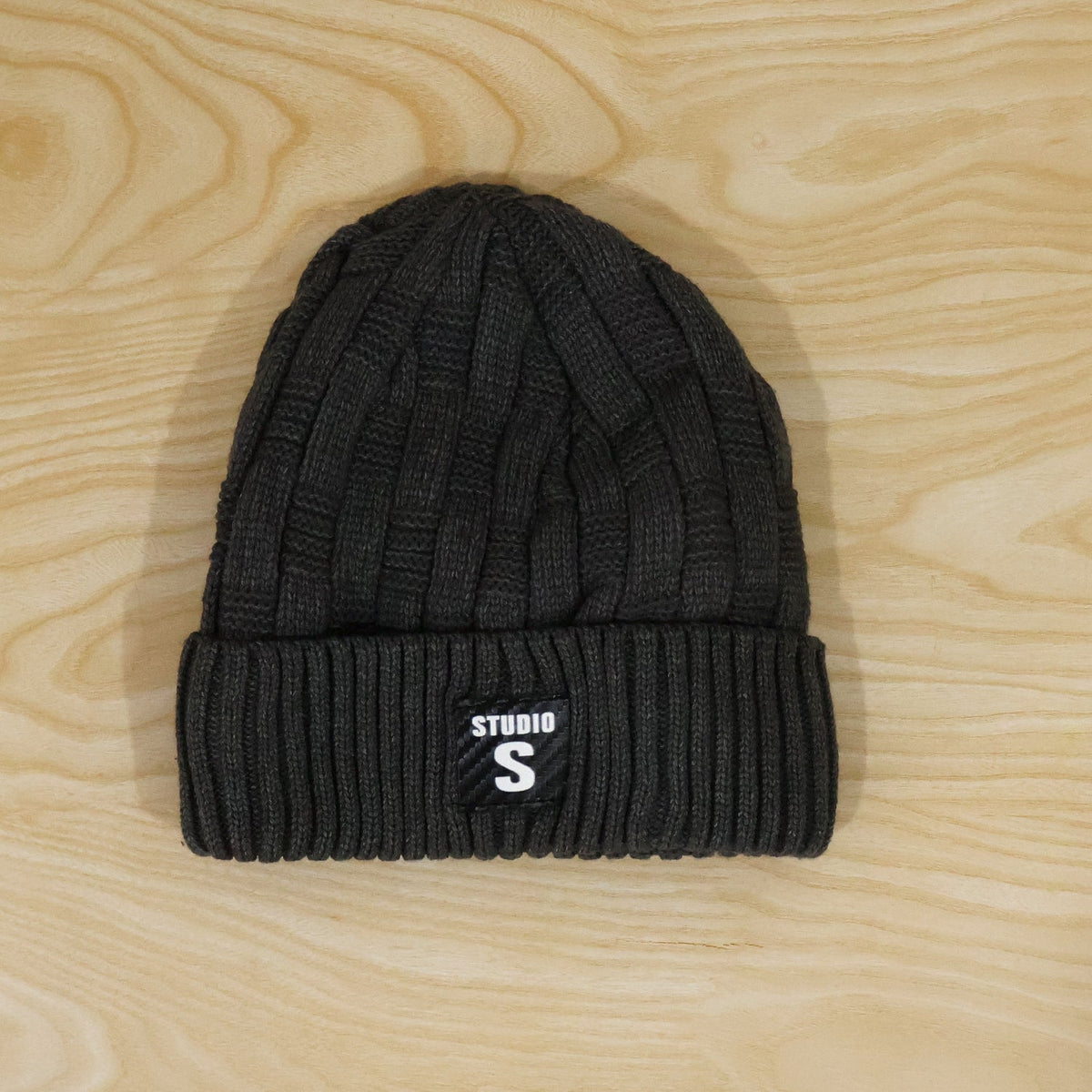 A dark grey cable knit beanie with a ribbed cuff and a black label with "STUDIO S" in white, lying flat on a light wooden