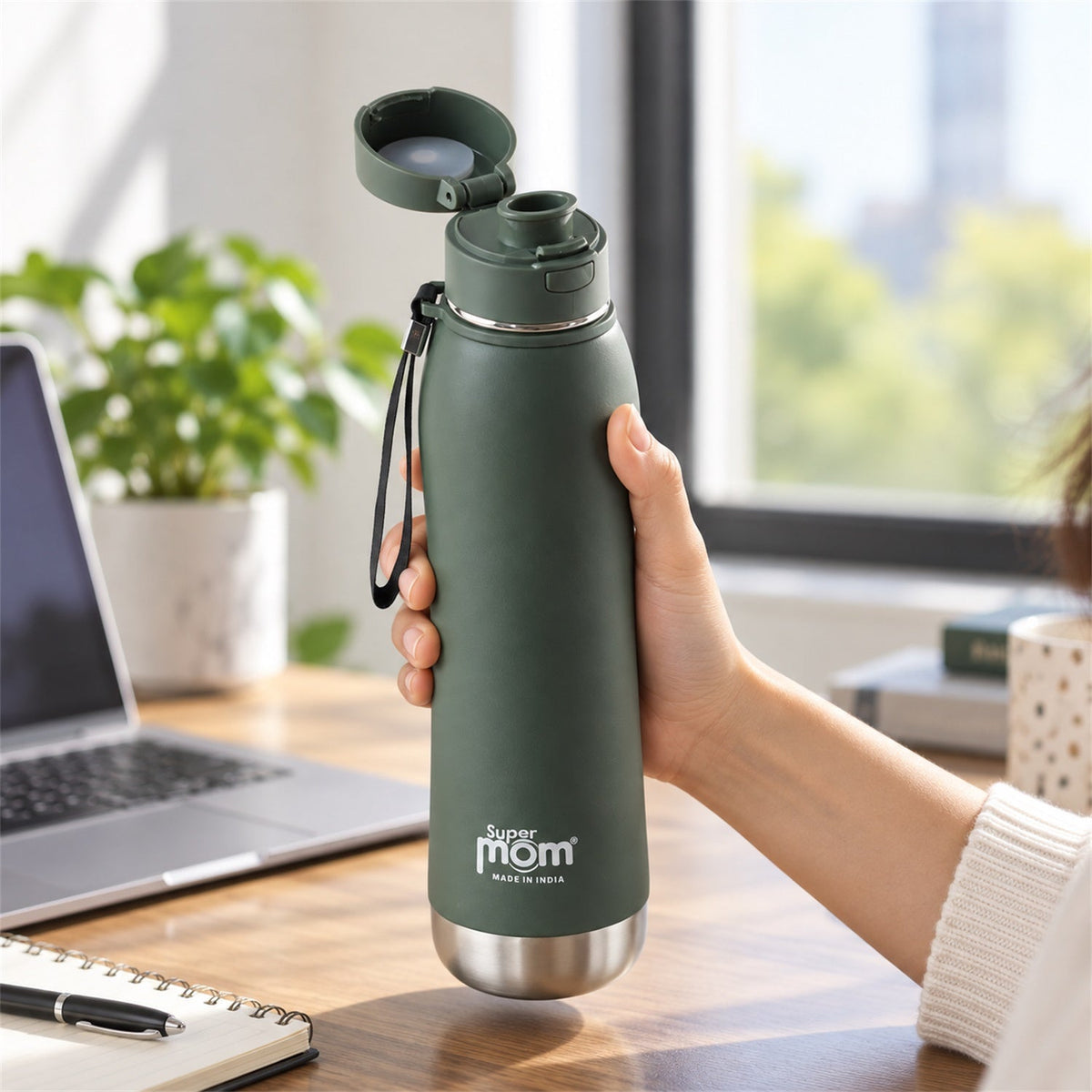 A person's hand holds an open dark green insulated stainless steel water bottle with a flip lid at a desk with a laptop and