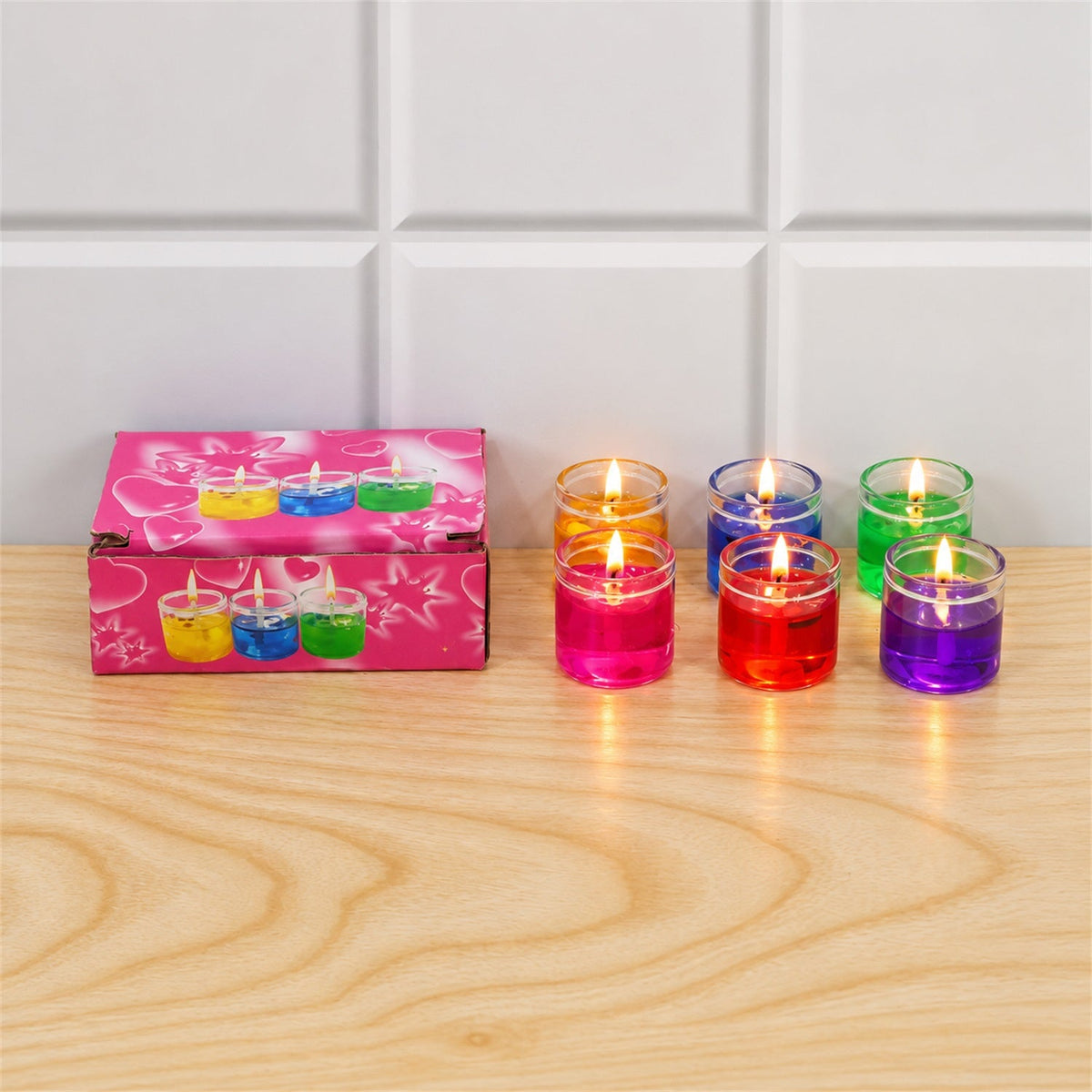 Six lit gel candles in glass jars, in yellow, blue, green, pink, red, and purple, arranged next to a pink box.