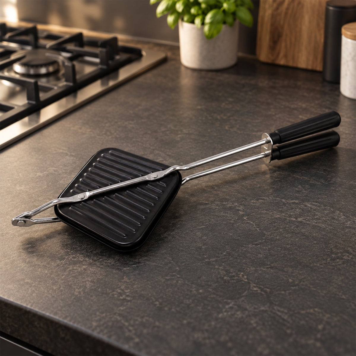Black stovetop sandwich maker with long silver handles resting on a dark grey speckled kitchen counter next to a gas stove.