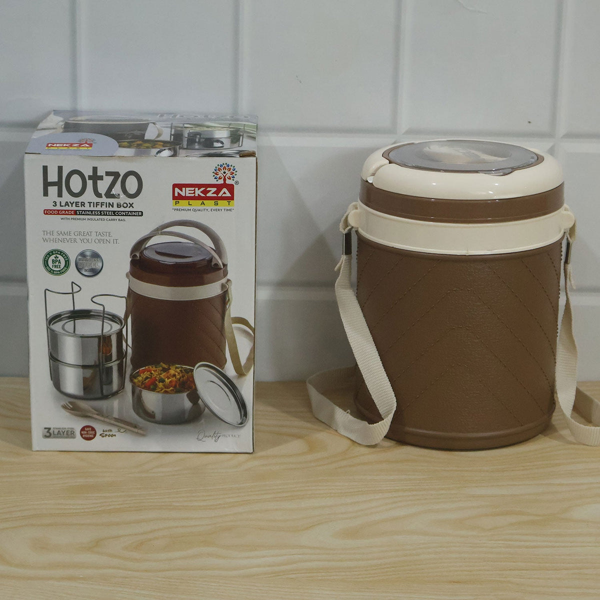 A brown and cream insulated tiffin box next to its product packaging, which displays the brand name "Hotzo Nekza" and an