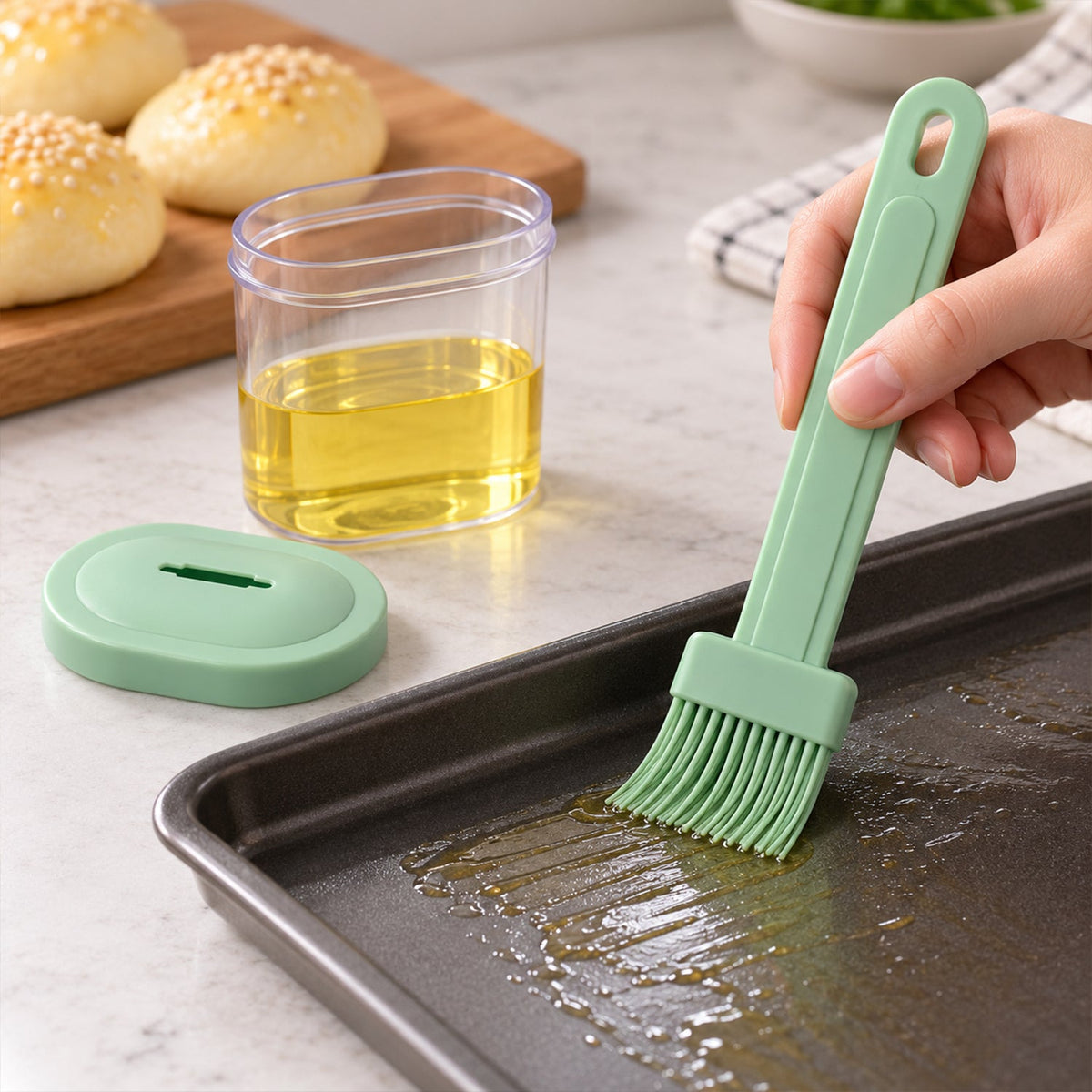 Hand holding a green silicone brush spreading oil on a dark baking sheet with three golden buns in the background.