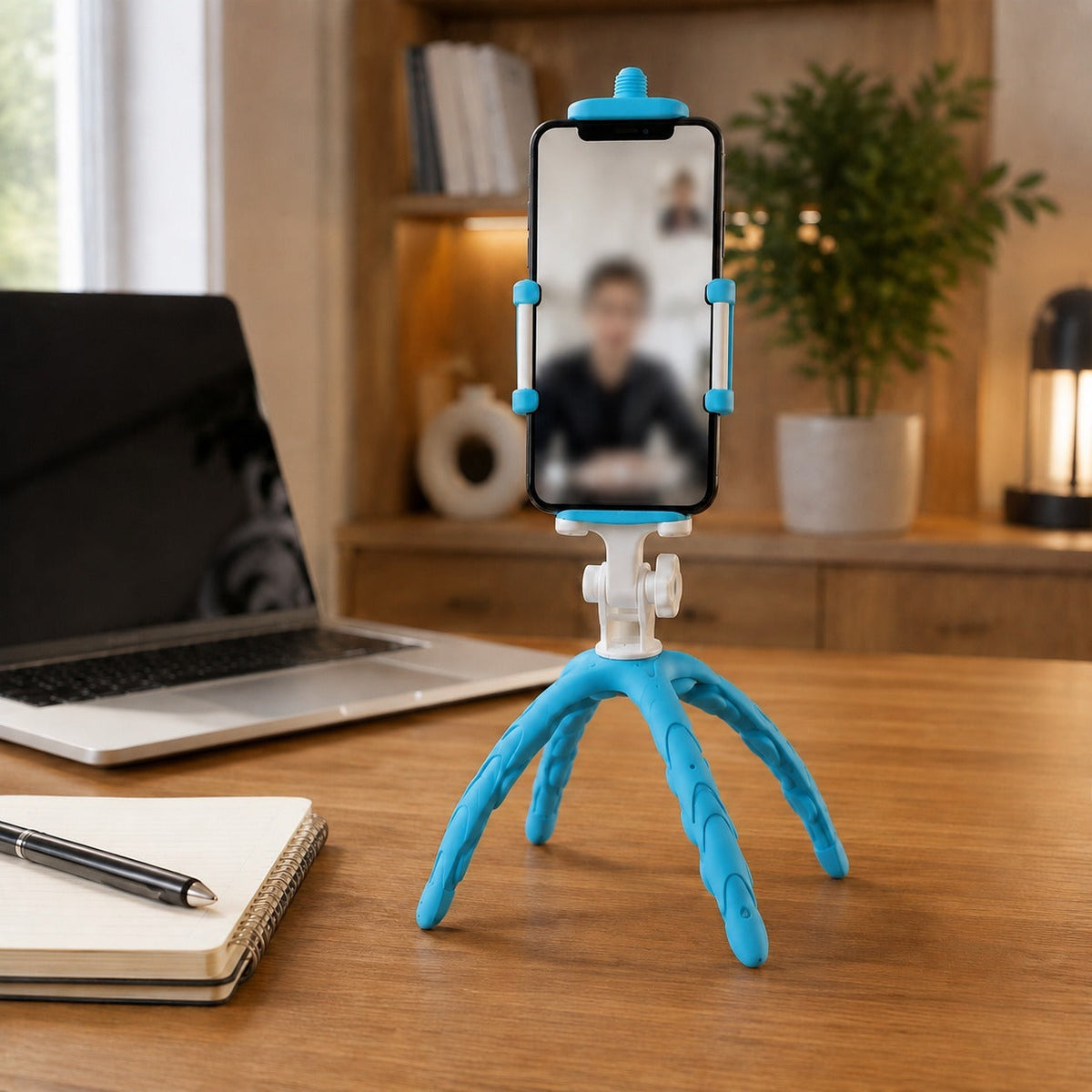 A blue and white flexible tripod holding a phone displaying a video call on a wooden desk next to a laptop and pen.