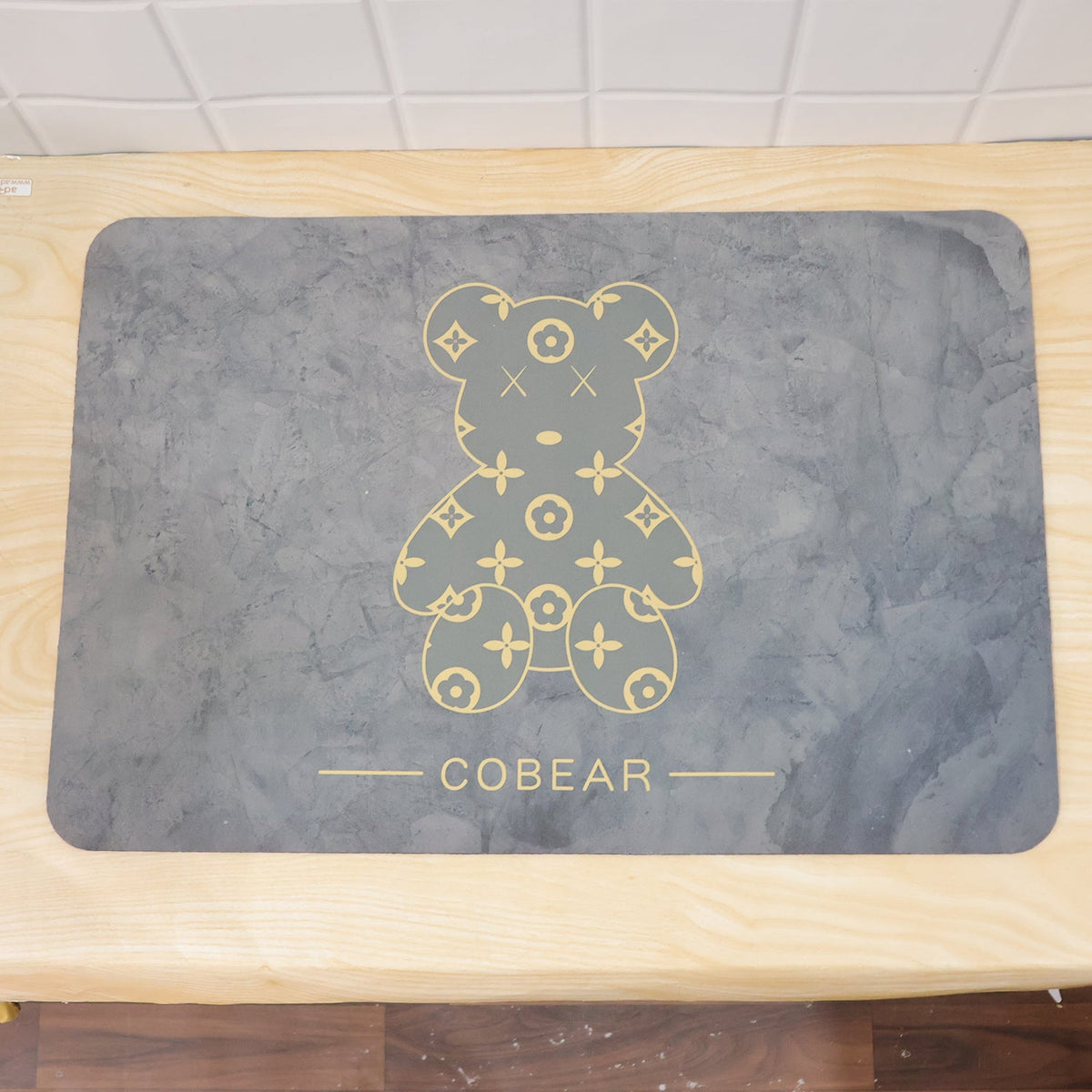 The grey rectangular bath mat with its golden bear design is displayed on a light wooden surface.