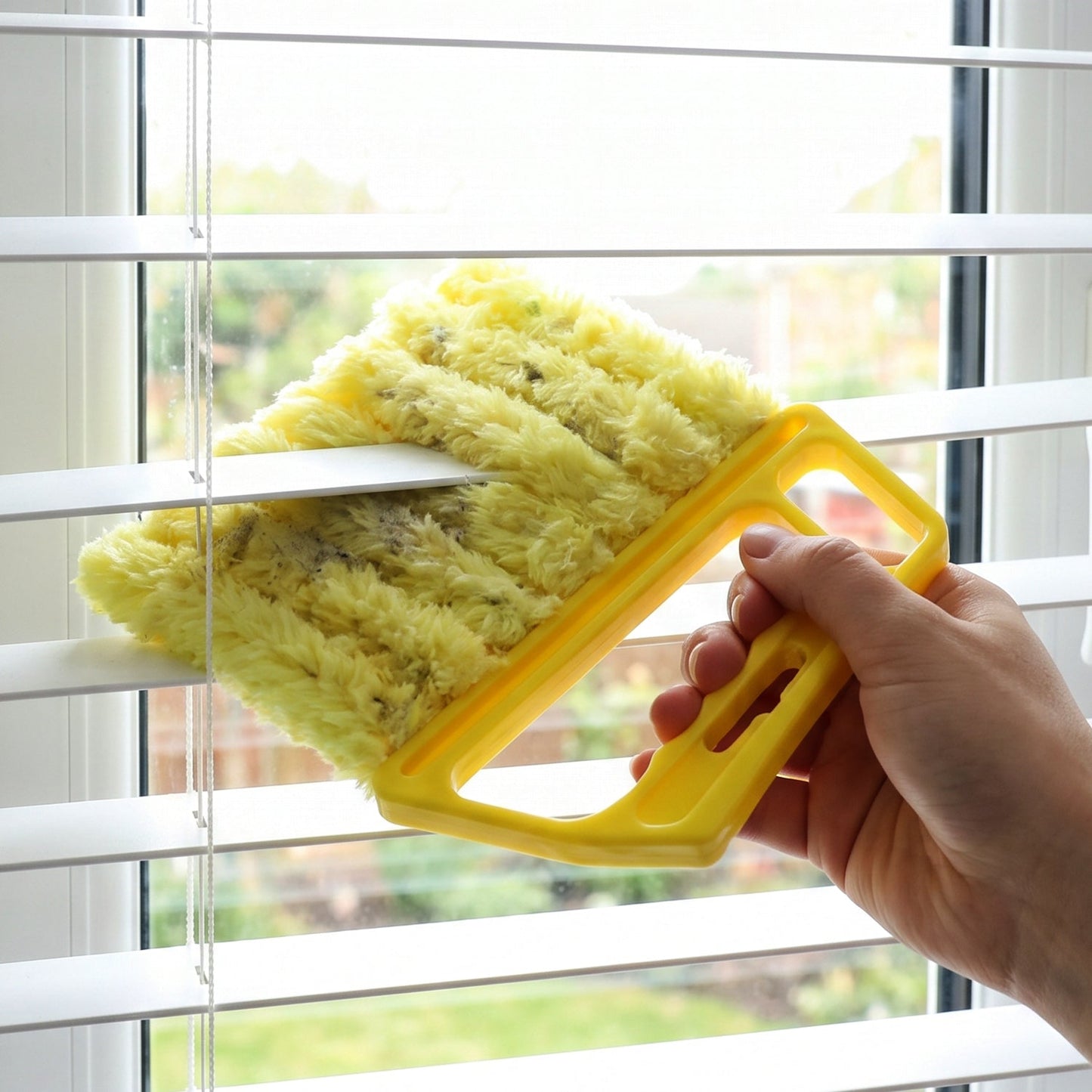A hand uses a yellow microfiber blind cleaner to wipe dust from white horizontal blinds, seen through a window.