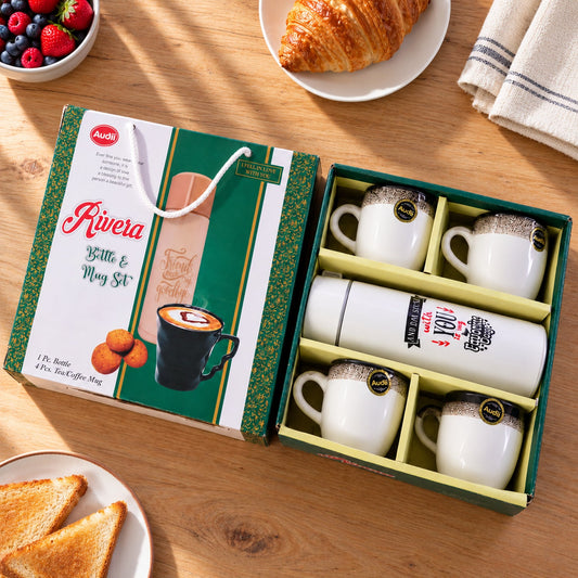 Open gift box revealing a white bottle and four white ceramic mugs with black speckled rims on a wooden table.