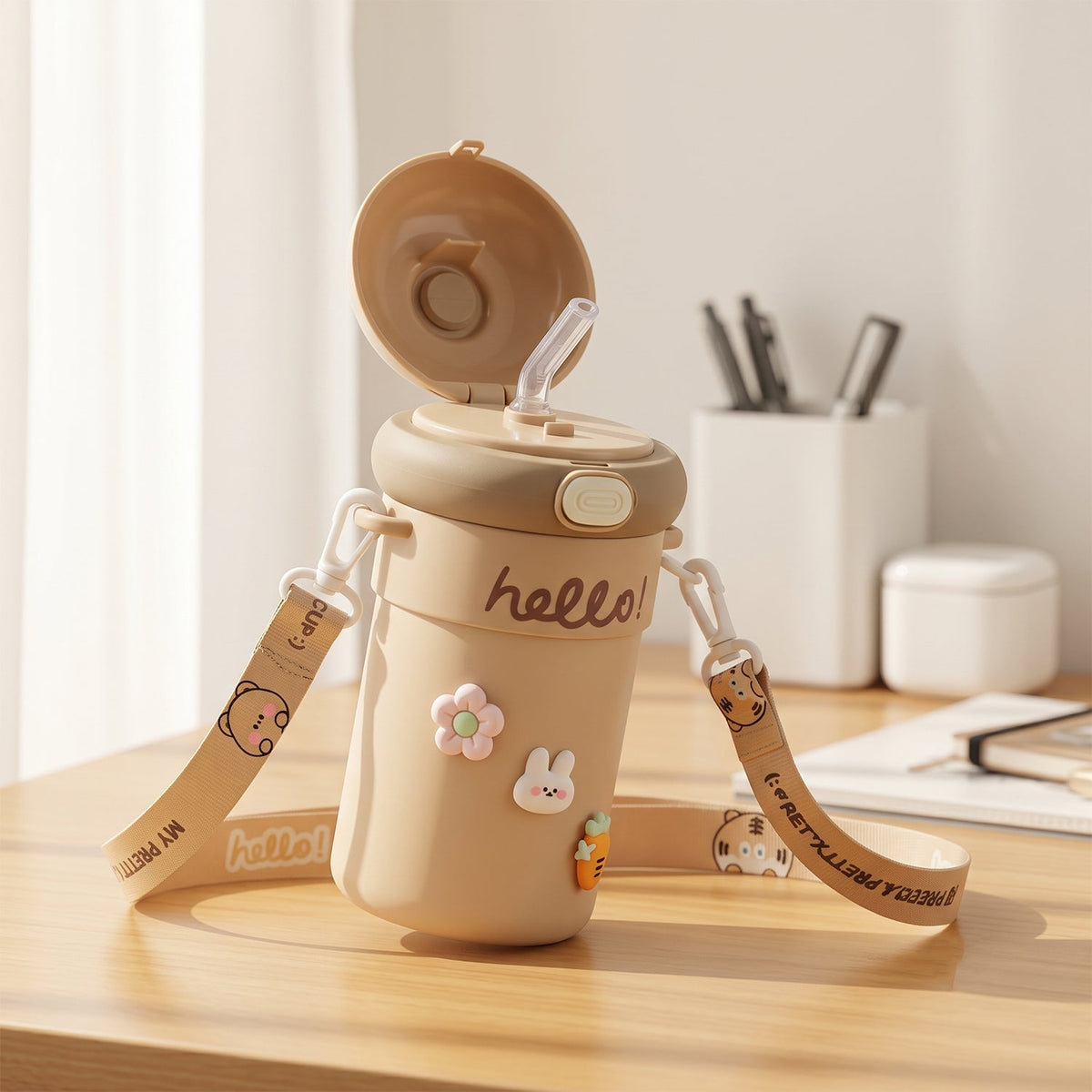 An open tan sipper bottle with a straw, 'hello!' inscription, and cute animal and flower charms, with a patterned strap, on