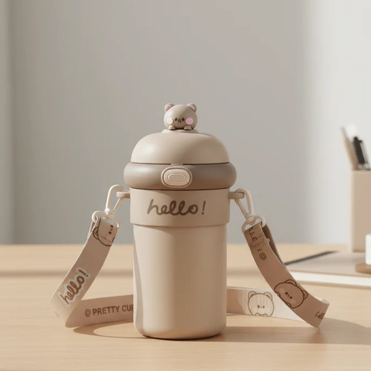 A tan sipper bottle with a bear on top, a 'hello!' inscription, and a patterned strap sits on a desk.