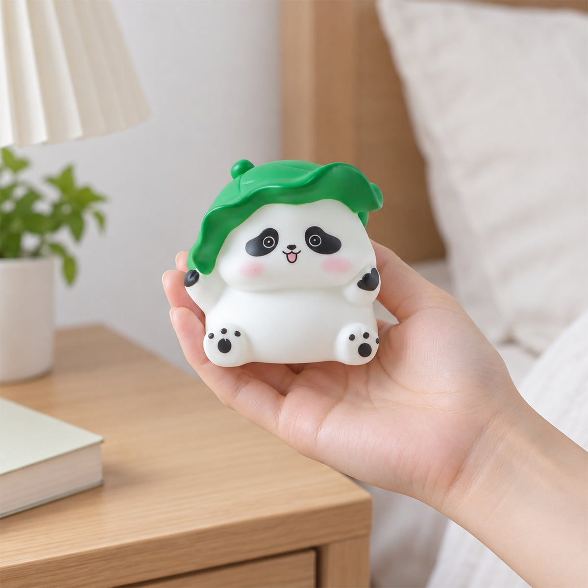 A hand holds the small, white panda night light with a green leaf hat, unlit, in a bedroom setting.