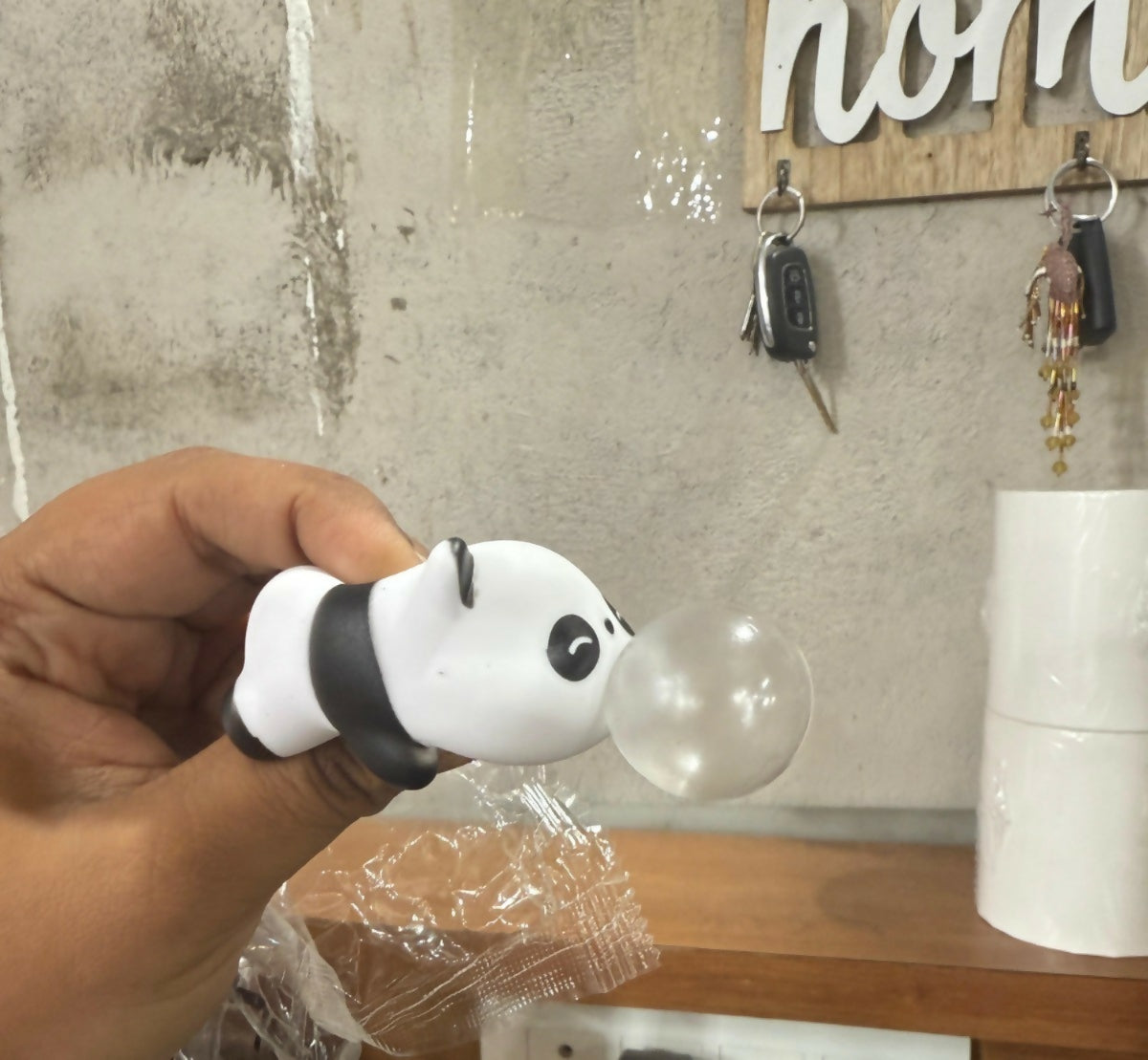A hand holds a white and black panda toy with a clear bubble, against a blurred background of a concrete wall and wooden key