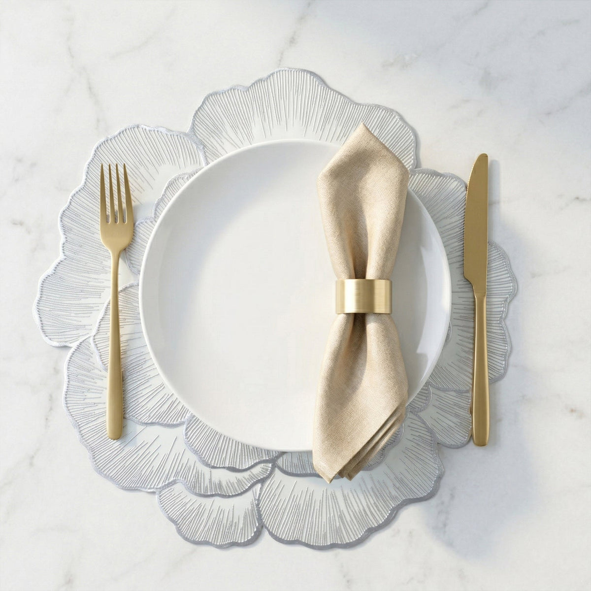 A silver floral placemat with a white plate, beige napkin, and golden cutlery on a marble surface.
