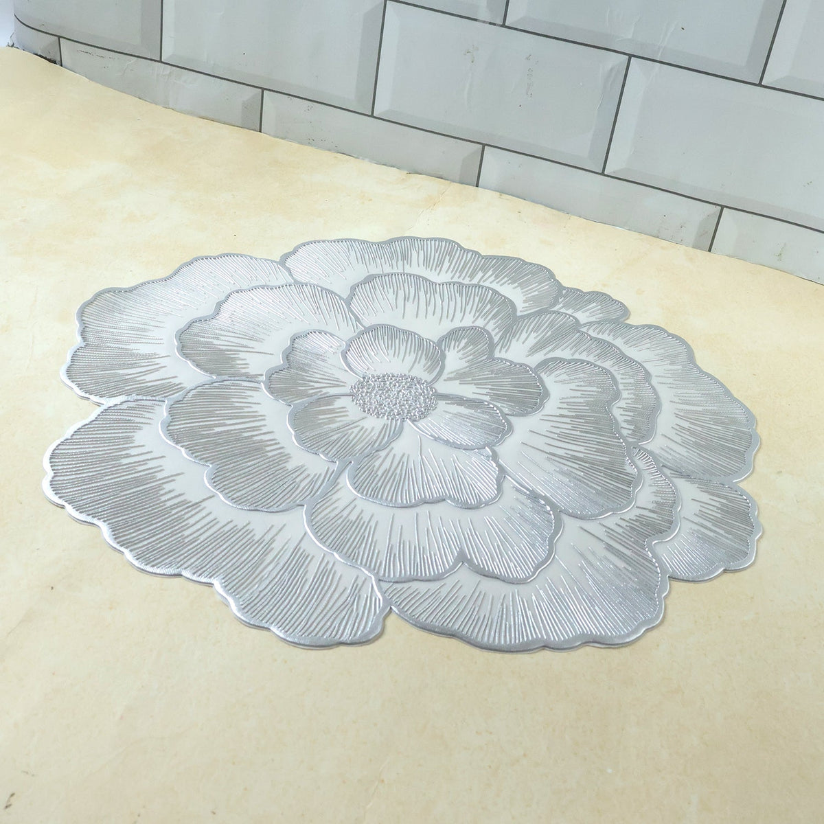A silver floral placemat with a textured design on a light brown surface with white tiled background.