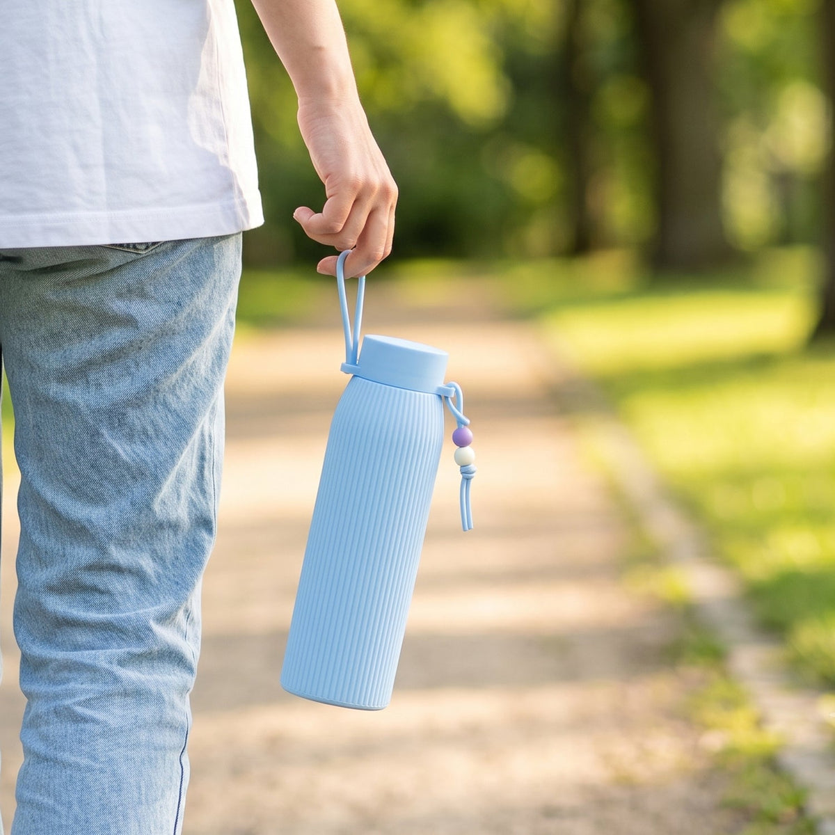 A person walks outdoors, holding a light blue ribbed glass water bottle with a beaded strap by their side.