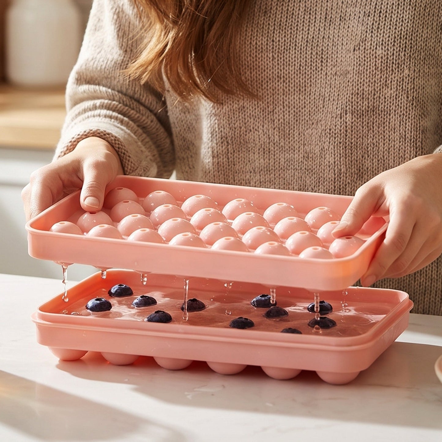 A person holds a pink ice ball tray, pouring water and blueberries into the bottom tray.