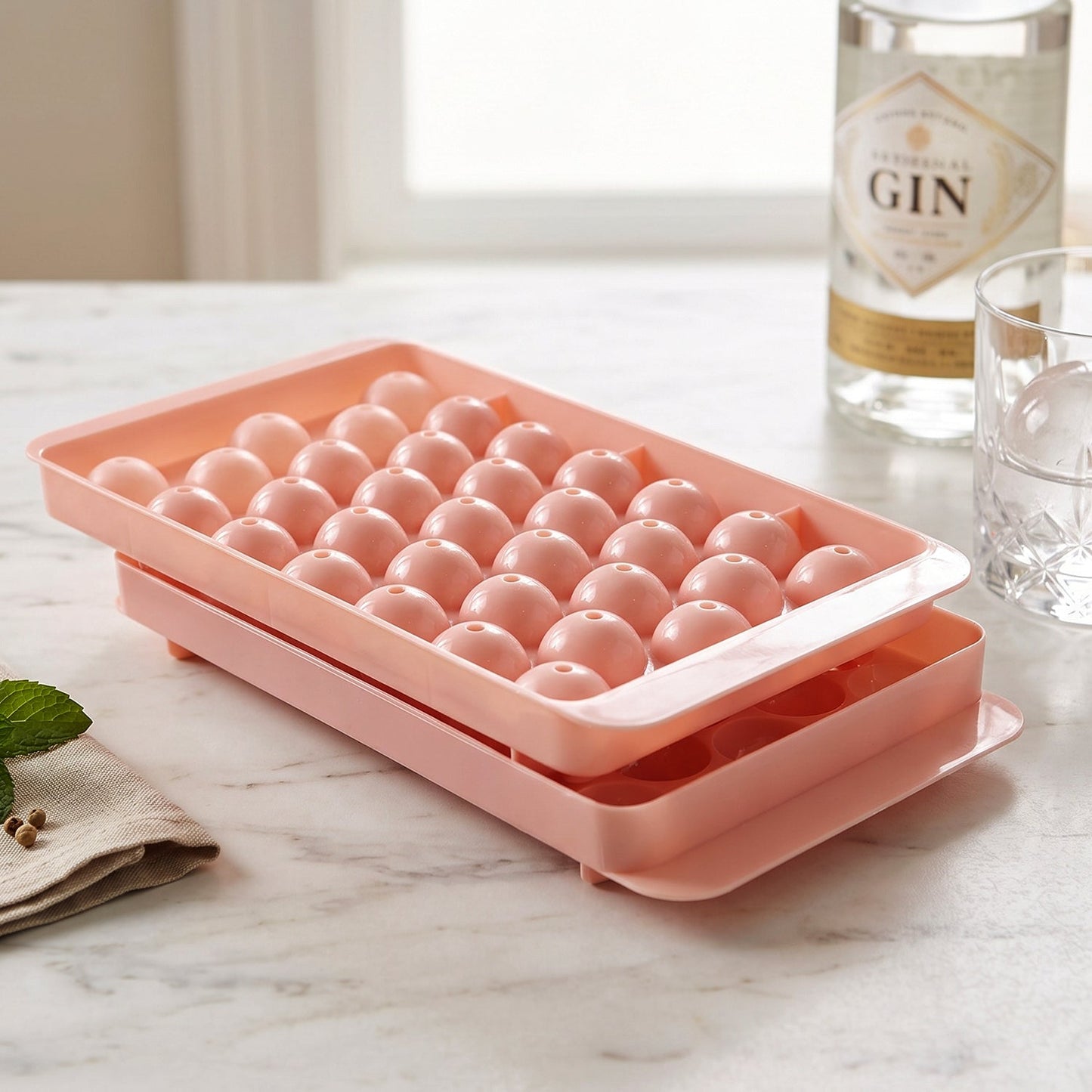 Two pink plastic ice ball trays stacked on a marble counter, with a bottle of gin and a glass in the background.
