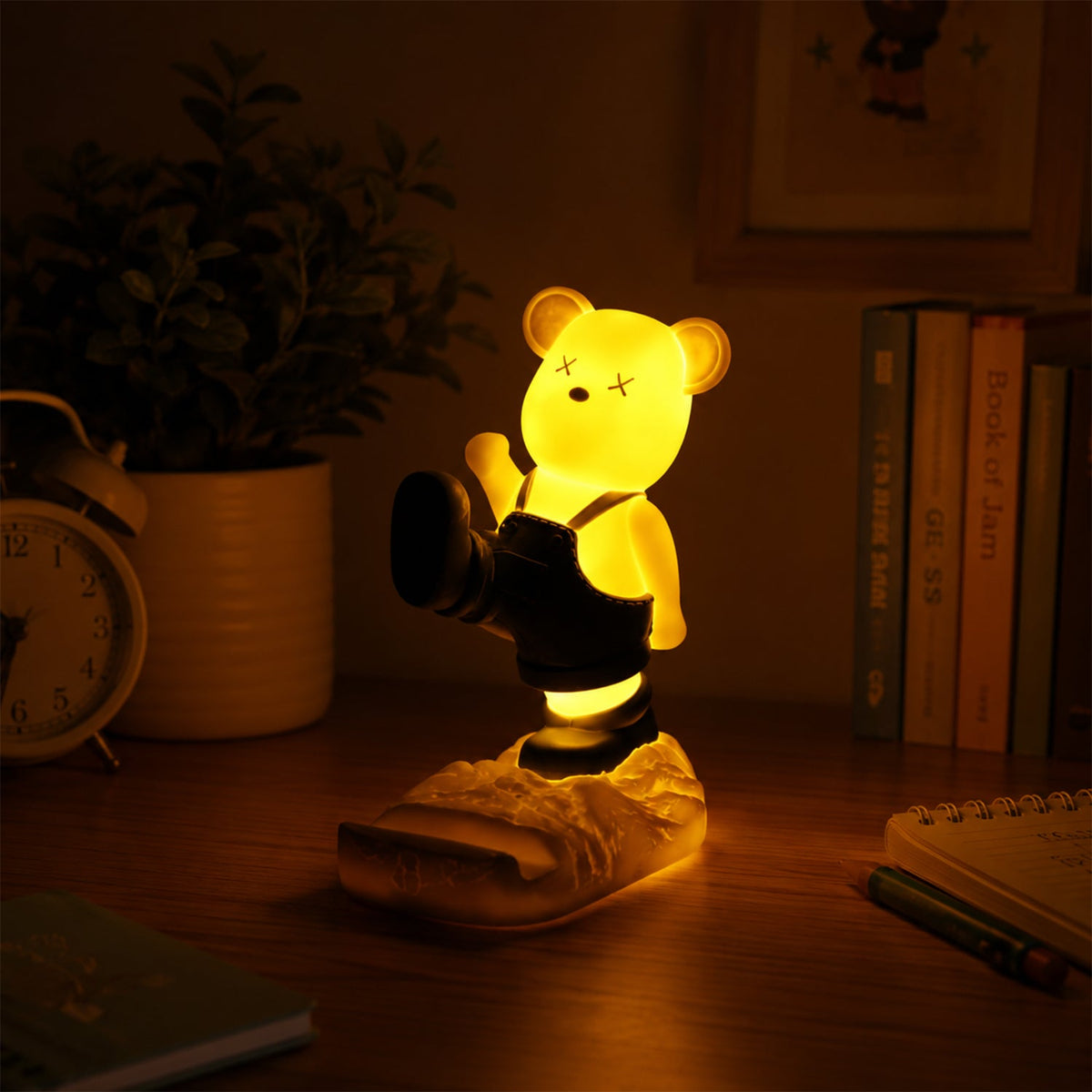 A glowing yellow bear night light and phone stand on a dark desk beside books and an alarm clock.