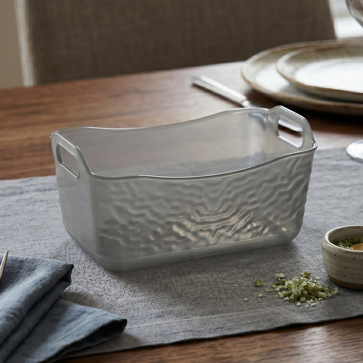 A rectangular dark gray textured glass storage basket with handles, placed on a light gray table runner with a small dish.