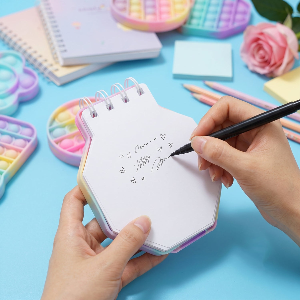 A person's hands writing on a pastel hexagon pop it fidget notebook with a black pen, surrounded by office supplies.