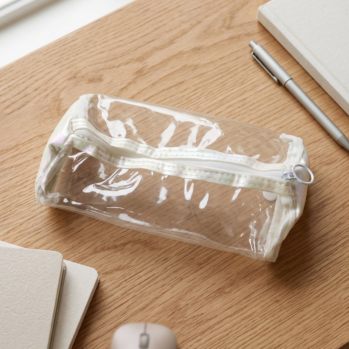 Empty clear PVC pencil case with white trim and zipper on a wooden desk next to a pen and notebook.