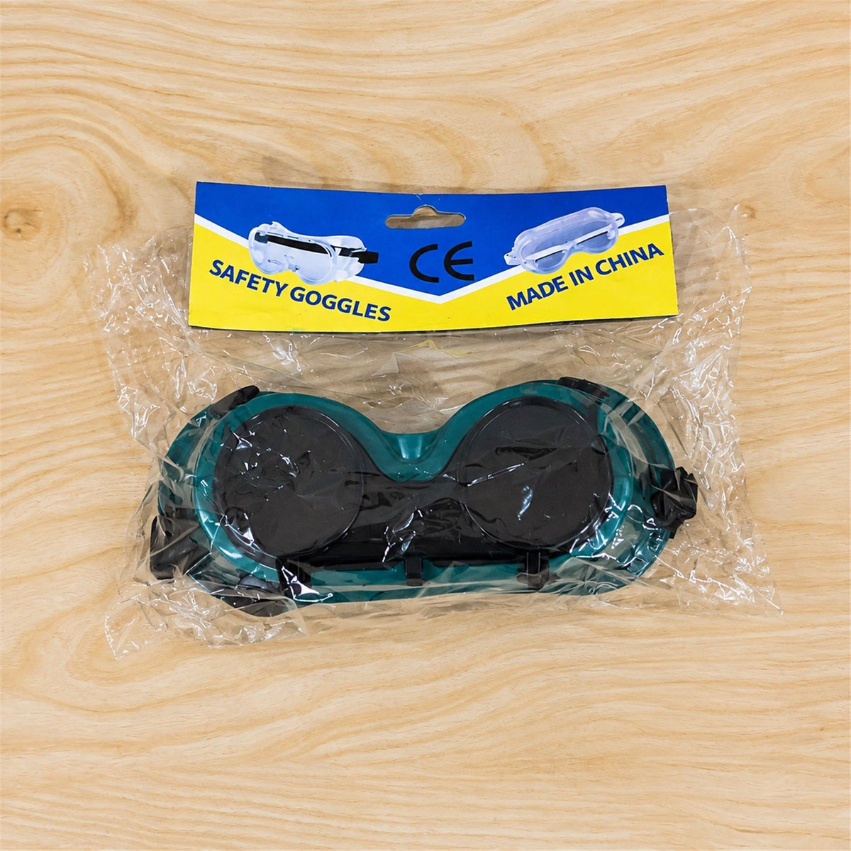 Green welding goggles in clear plastic packaging with a blue and yellow header card on a wooden surface.