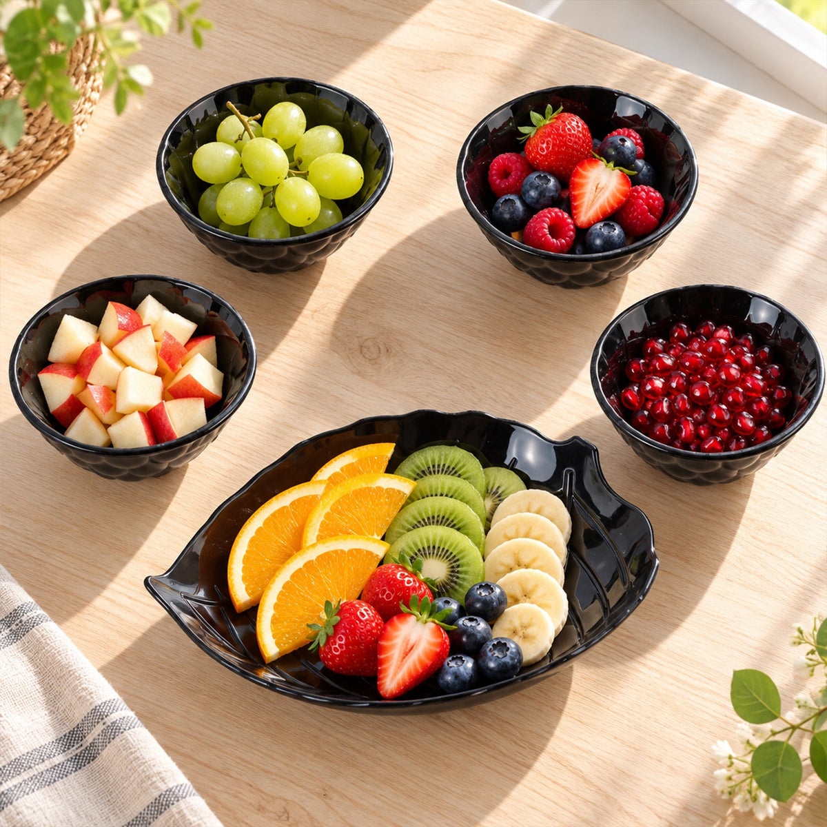 A black leaf-shaped platter holds sliced fruit, with four small black bowls filled with different fresh fruits.