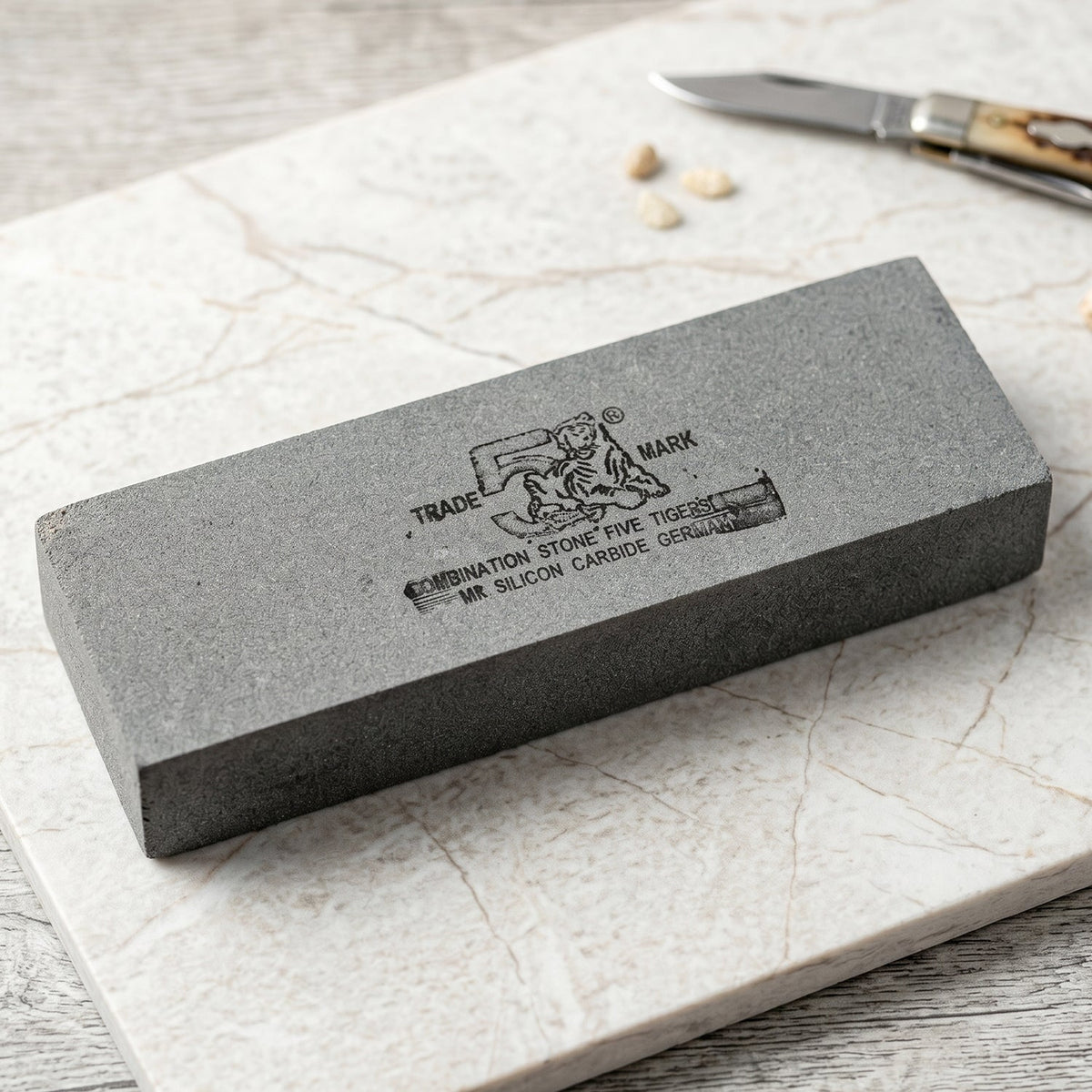 Gray rectangular sharpening stone with black text and tiger logo on a white marble surface next to a small knife.