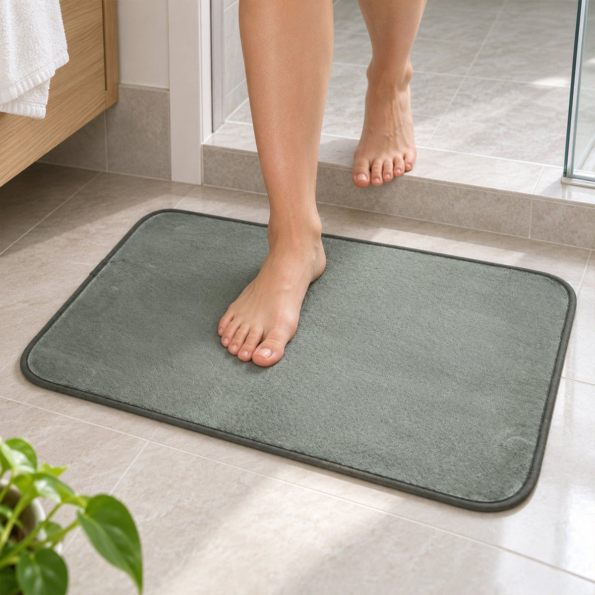 Bare feet stepping onto a soft, grey floor mat placed on light bathroom tiles.
