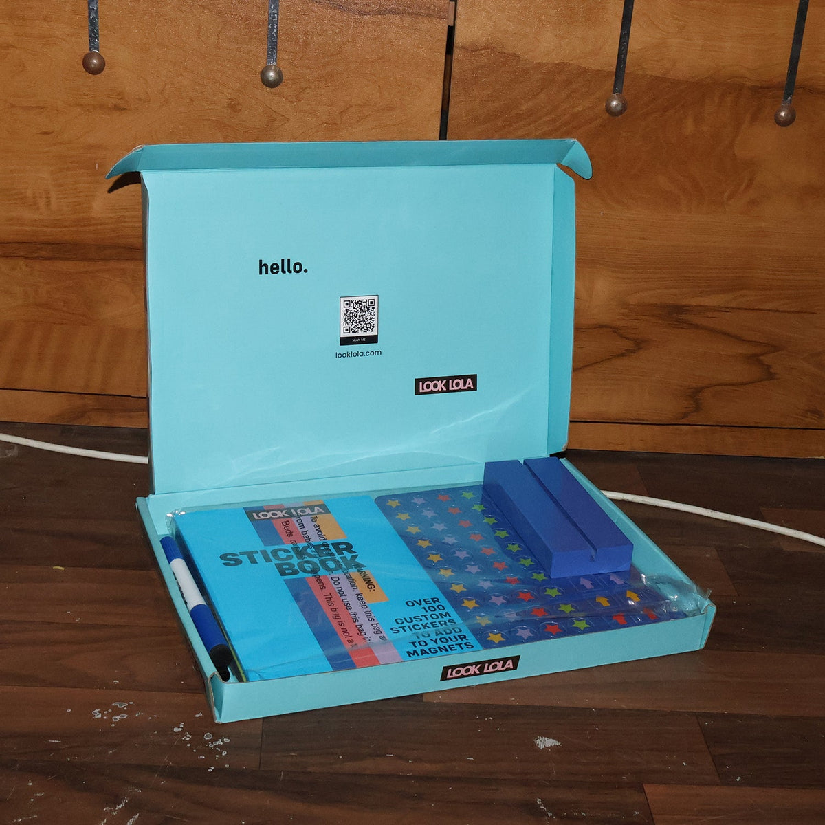 An open light blue box containing a sticker book, blue magnetic strips, and a pen, on a wooden floor.