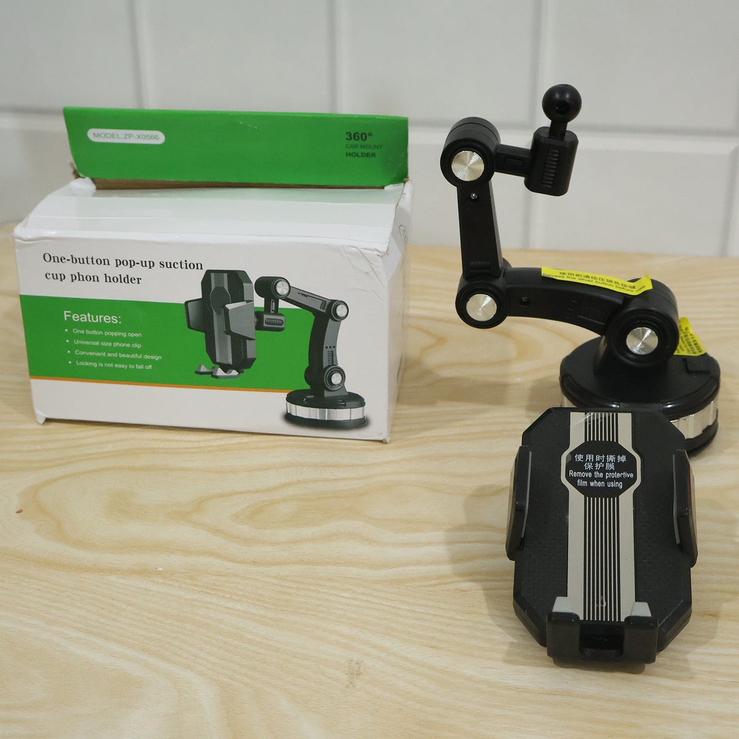 A black car mount phone holder is disassembled next to its white and green packaging box on a light wooden surface.