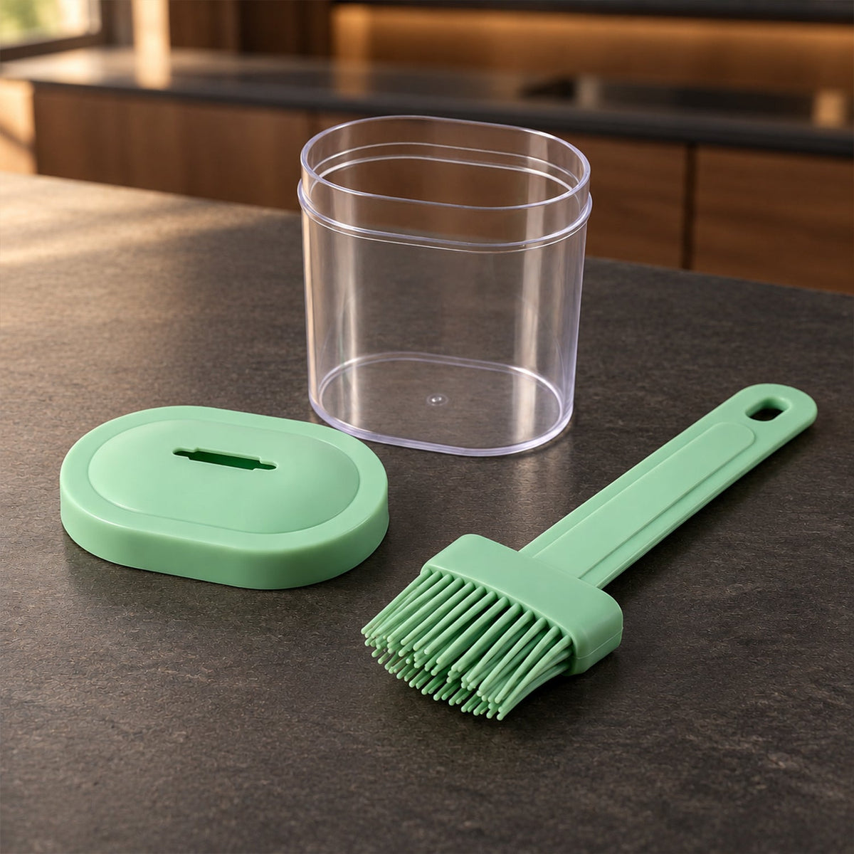 A green silicone basting brush, a clear empty container, and a green oval lid with a slot are arranged on a dark countertop.
